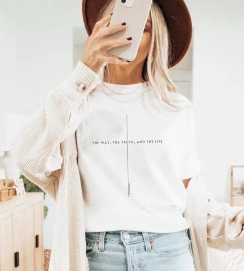 May include: A white short-sleeved t-shirt featuring a thin vertical black line forming a cross shape with the text "THE WAY, THE TRUTH, AND THE LIFE" printed horizontally across the center. The shirt is worn with a beige textured cardigan and blue jeans.