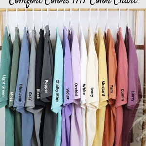 May include: A collection of Comfort Colors 1717 t-shirts hanging on a wooden rack, displayed in a spectrum of colors including Light Green, Moss, Blue Jean, Gray, Black, Pepper, Chalky Mint, Violet, Orchid, White, Ivory, Mustard, Yam, Cumin, and Berry.