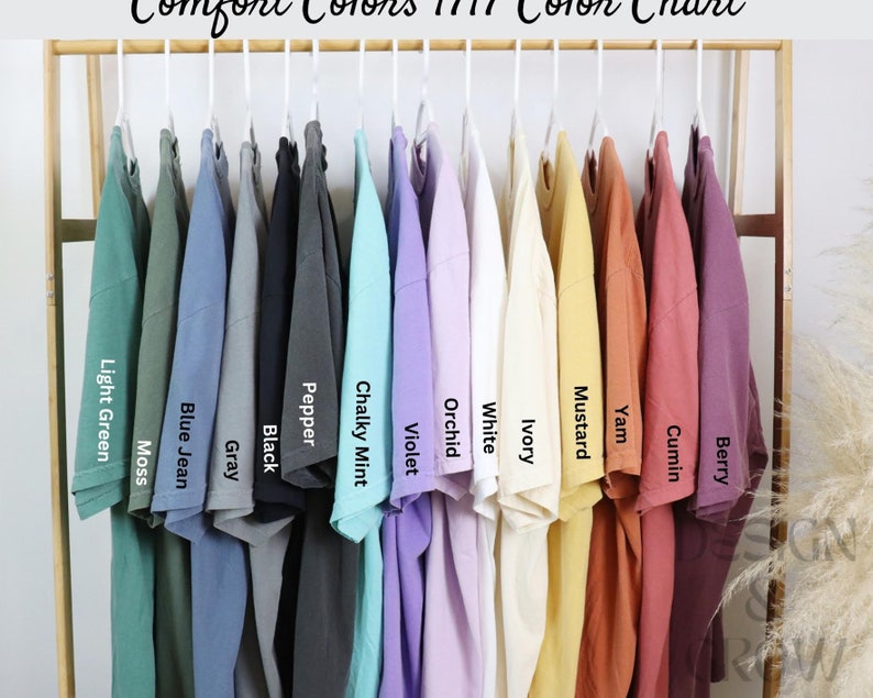 May include: A collection of Comfort Colors 1717 t-shirts hanging on a wooden rack, displayed in a spectrum of colors including Light Green, Moss, Blue Jean, Gray, Black, Pepper, Chalky Mint, Violet, Orchid, White, Ivory, Mustard, Yam, Cumin, and Berry.