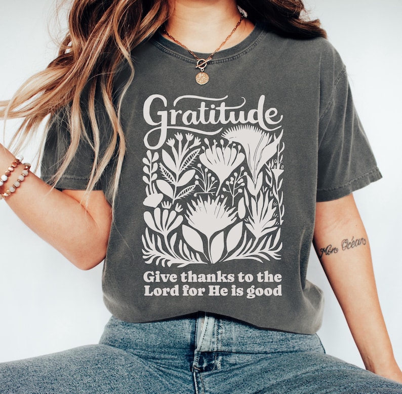May include: A charcoal gray short-sleeve t-shirt featuring a cream-colored graphic print. The design includes the word Gratitude above a collection of stylized floral illustrations and the text Give thanks to the Lord for He is good.