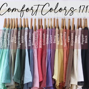 May include: A rack of 20 t-shirts in various colours, hanging on wooden hangers. The t-shirts are labelled with names such as "Chalky Mint", "Island Reef", "Seafoam", "Bay", "Khaki", "Light Green", "Moss", "Chambray", "Blue Jean", "Blossom", "Crunchberry", "Watermelon", "Crimson", "Orchid", "Violet", "Berry", "Banana", "Mustard", "Terracotta", "Yam", "Espresso", "Ivory", "White", "Pepper", and "Black". The text "Comfort Colors 1717" is displayed at the top of the image.