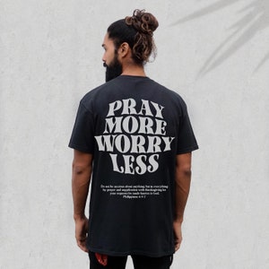Puede incluir: Camiseta negra con texto blanco que dice "Reza m&aacute;s, preoc&uacute;pate menos". El texto est&aacute; en un estilo de fuente retro. Debajo del texto hay una cita de Filipenses 4:6-7.