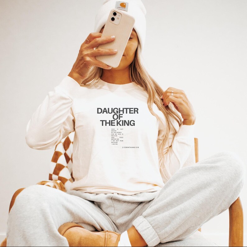 May include: White long-sleeved shirt with the text "Daughter of the King" and a list of Bible verses. 2 Corinthians 6:18.