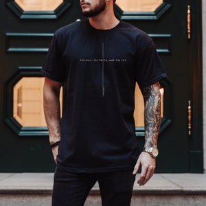 May include: A black short-sleeved t-shirt featuring a vertical white line design centered on the chest. The text "THE WAY, THE TRUTH, AND THE LIFE" is printed in white capital letters across the chest, with "JOHN 14:6" written vertically below.