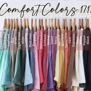 May include: A collection of Comfort Colors 1717 t-shirts hanging on a gold rack. The shirts are displayed in a spectrum of colors including Chalky Mint, Island Reef, Seafoam, Bay, Khaki, Light Green, Moss, Chambray, Blue Jean, Blossom, Crunchberry, Watermelon, Crimson, Orchid, Violet, Berry, Banana, Mustard, Terracotta, Yam, Espresso, Ivory, White, Pepper, and Black.