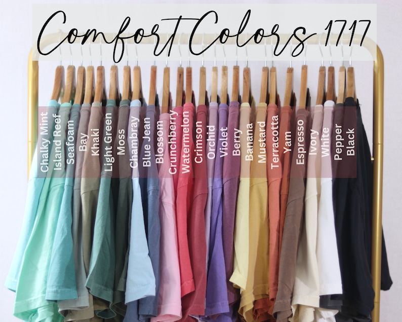 May include: A collection of Comfort Colors 1717 t-shirts hanging on a gold rack. The shirts are displayed in a spectrum of colors including Chalky Mint, Island Reef, Seafoam, Bay, Khaki, Light Green, Moss, Chambray, Blue Jean, Blossom, Crunchberry, Watermelon, Crimson, Orchid, Violet, Berry, Banana, Mustard, Terracotta, Yam, Espresso, Ivory, White, Pepper, and Black.