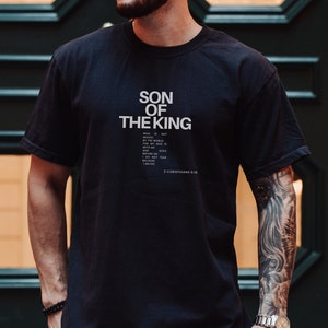 May include: A black short-sleeved t-shirt featuring the white text SON OF THE KING printed in a bold, sans-serif font. Below this, smaller white text reads: WHO IS NOT MOVED BY THE WORLD. FOR MY GOD IS WITH ME AND GOES BEFORE ME. 2 CORINTHIANS 6:18.