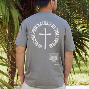 Pode incluir: Uma camiseta cinza com um gr&aacute;fico circular branco nas costas. O gr&aacute;fico apresenta uma cruz e o texto "No weapon formed against me shall prosper." O texto "And every tongue that rises against thee in judgment thou shalt condemn. This is the heritage of the servants of the Lord, and their righteousness is from me, says the Lord." est&aacute; impresso abaixo do gr&aacute;fico.