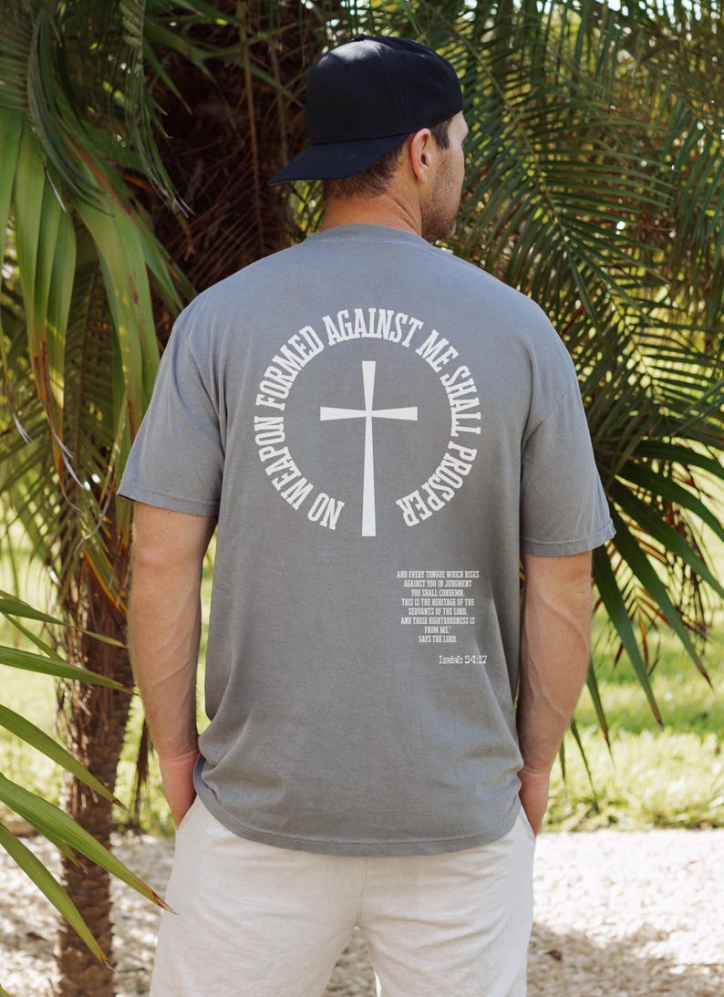 Pode incluir: Uma camiseta cinza com um gr&aacute;fico circular branco nas costas. O gr&aacute;fico apresenta uma cruz e o texto "No weapon formed against me shall prosper." O texto "And every tongue that rises against thee in judgment thou shalt condemn. This is the heritage of the servants of the Lord, and their righteousness is from me, says the Lord." est&aacute; impresso abaixo do gr&aacute;fico.
