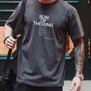 May include: A charcoal grey short-sleeve t-shirt featuring the white text SON OF THE KING followed by a smaller block of text that reads WHO IS NOT MOVED BY THE WORLD. FOR MY GOD IS WITH ME AND GOES BEFORE ME. I DO NOT FEAR BECAUSE I AM HIS. 2 CORINTHIANS 6:18.