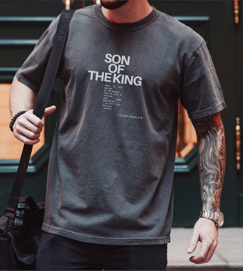 May include: A charcoal grey short-sleeve t-shirt featuring the white text SON OF THE KING followed by a smaller block of text that reads WHO IS NOT MOVED BY THE WORLD. FOR MY GOD IS WITH ME AND GOES BEFORE ME. I DO NOT FEAR BECAUSE I AM HIS. 2 CORINTHIANS 6:18.