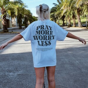 Puede incluir: Una camiseta azul claro con el texto "Pray More Worry Less" impreso en negro. La camiseta tambi&eacute;n incluye un vers&iacute;culo b&iacute;blico de Filipenses 4:6-7.