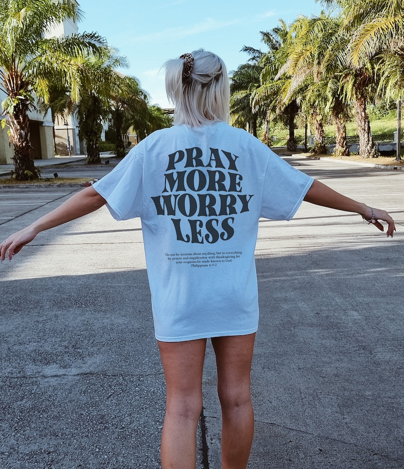 Puede incluir: Una camiseta azul claro con el texto "Pray More Worry Less" impreso en negro. La camiseta tambi&eacute;n incluye un vers&iacute;culo b&iacute;blico de Filipenses 4:6-7.