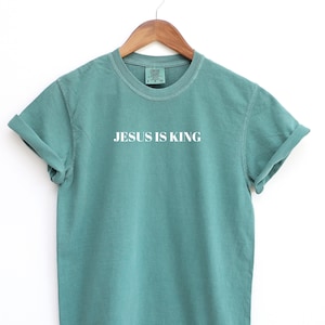 K&ouml;nnte beinhalten: Ein blaugr&uuml;nes T-Shirt mit dem Aufdruck "JESUS IS KING" in Wei&szlig; auf der Vorderseite.