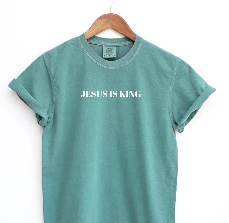 K&ouml;nnte beinhalten: Ein blaugr&uuml;nes T-Shirt mit dem Aufdruck "JESUS IS KING" in Wei&szlig; auf der Vorderseite.