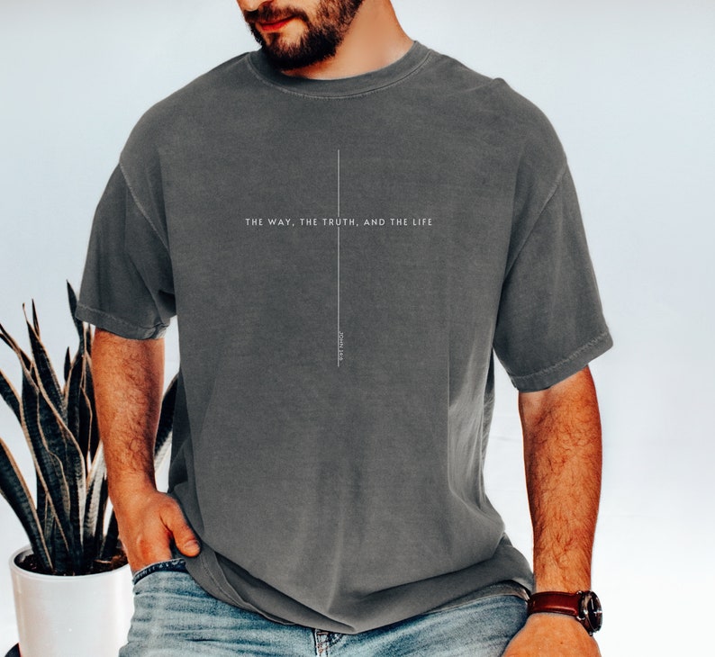 May include: A charcoal gray short-sleeve t-shirt featuring a thin white vertical line forming a cross shape. The text "THE WAY, THE TRUTH, AND THE LIFE" is printed horizontally across the center, with "JOHN 14:6" written vertically below it.