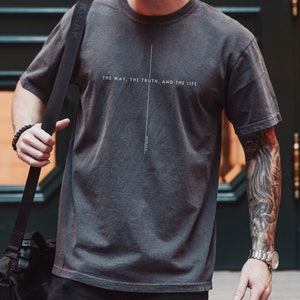 May include: A charcoal gray short-sleeve t-shirt featuring a thin vertical white line forming a cross shape. The text "THE WAY, THE TRUTH, AND THE LIFE" is printed horizontally across the center, with "JOHN 14:6" printed vertically below the cross.