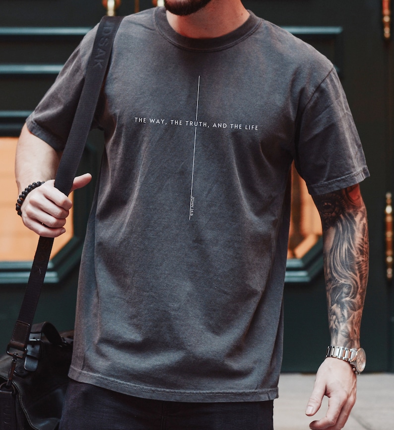 May include: A charcoal gray short-sleeve t-shirt featuring a thin vertical white line forming a cross shape. The text "THE WAY, THE TRUTH, AND THE LIFE" is printed horizontally across the center, with "JOHN 14:6" printed vertically below the cross.