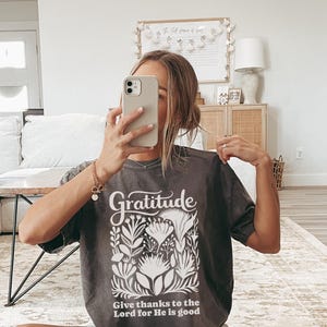 May include: A charcoal grey short-sleeve t-shirt featuring the word Gratitude in a white script font above a white botanical illustration and the text Give thanks to the Lord for He is good printed in a white sans-serif font at the bottom.