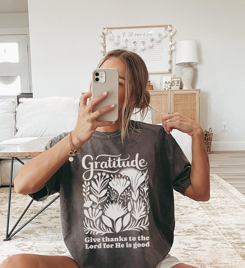 May include: A charcoal grey short-sleeve t-shirt featuring the word Gratitude in a white script font above a white botanical illustration and the text Give thanks to the Lord for He is good printed in a white sans-serif font at the bottom.