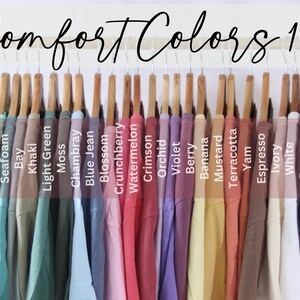 May include: A collection of Comfort Colors 1717 t-shirts hanging on a gold rack, displayed in a spectrum of colors including Chalky Mint, Island Reef, Seafoam, Bay, Khaki, Light Green, Moss, Chambray, Blue Jean, Blossom, Crunchberry, Watermelon, Crimson, Orchid, Violet, Berry, Banana, Mustard, Terracotta, Yam, Espresso, Ivory, White, Pepper, and Black.