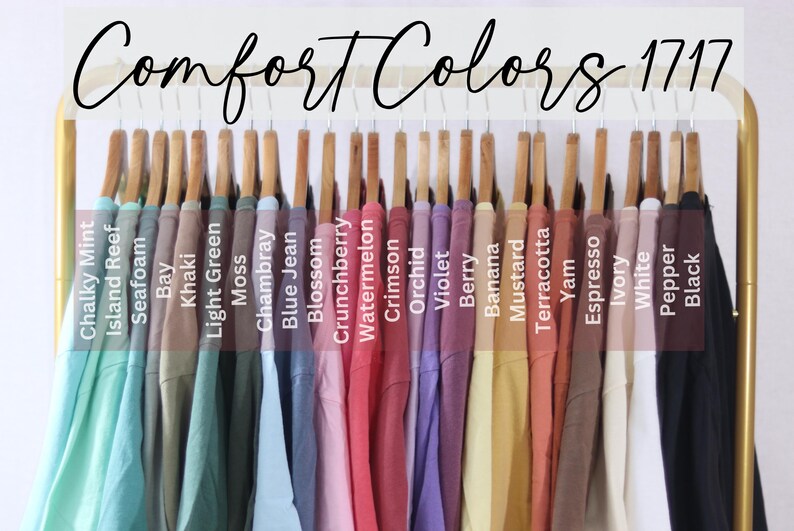 May include: A collection of Comfort Colors 1717 t-shirts hanging on a gold rack, displayed in a spectrum of colors including Chalky Mint, Island Reef, Seafoam, Bay, Khaki, Light Green, Moss, Chambray, Blue Jean, Blossom, Crunchberry, Watermelon, Crimson, Orchid, Violet, Berry, Banana, Mustard, Terracotta, Yam, Espresso, Ivory, White, Pepper, and Black.
