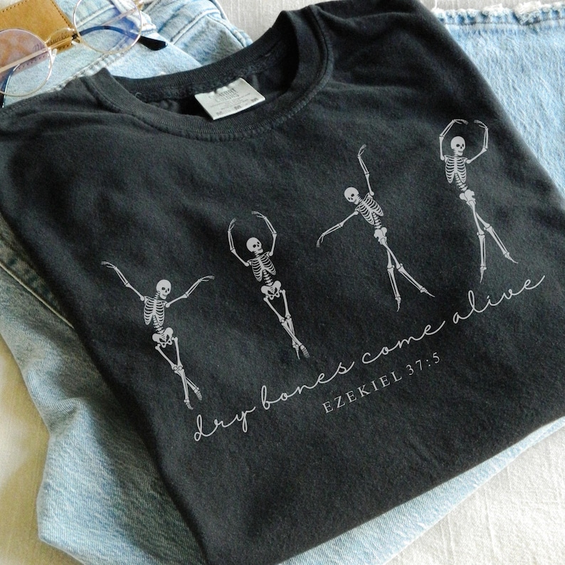 May include: Black t-shirt with a white graphic of four skeletons dancing. The text "dry bones come alive" and "EZEKIEL 37:5" is printed below the skeletons.