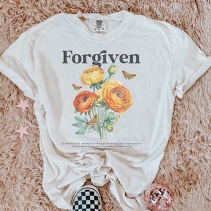 May include: A white t-shirt with the word "Forgiven" printed in black. The shirt features a floral design with yellow and orange flowers and butterflies. The text "In Him we have redemption through His blood, the forgiveness of sins in accordance with the riches of God's grace Ephesians 1:7" is printed below the floral design.