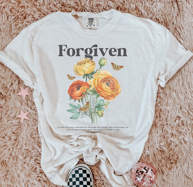 May include: A white t-shirt with the word "Forgiven" printed in black. The shirt features a floral design with yellow and orange flowers and butterflies. The text "In Him we have redemption through His blood, the forgiveness of sins in accordance with the riches of God's grace Ephesians 1:7" is printed below the floral design.