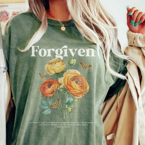 May include: A green t-shirt with the word "Forgiven" printed in white. The shirt features a floral design with yellow and orange flowers and butterflies. The text "In Him we have redemption through His blood, the forgiveness of sins in accordance with the riches of God's grace Ephesians 1:7" is printed below the floral design.