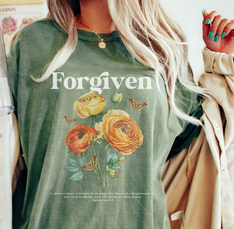 May include: A green t-shirt with the word "Forgiven" printed in white. The shirt features a floral design with yellow and orange flowers and butterflies. The text "In Him we have redemption through His blood, the forgiveness of sins in accordance with the riches of God's grace Ephesians 1:7" is printed below the floral design.