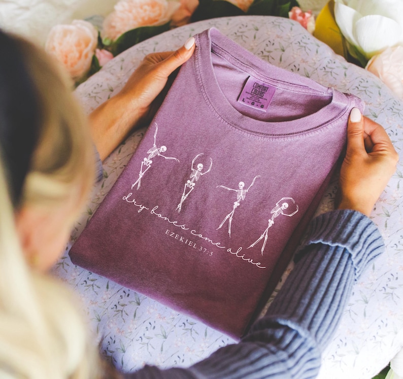 May include: Purple t-shirt with a white graphic of skeletons dancing and the text "dry bones come alive Ezekiel 37:3"