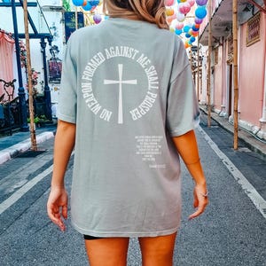 Pode incluir: Uma camiseta verde claro de tamanho grande com uma estampa gr&aacute;fica branca nas costas. O gr&aacute;fico apresenta uma cruz dentro de um c&iacute;rculo com o texto "No weapon formed against me shall prosper".