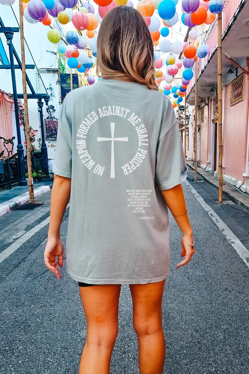 Pode incluir: Uma camiseta verde claro de tamanho grande com uma estampa gr&aacute;fica branca nas costas. O gr&aacute;fico apresenta uma cruz dentro de um c&iacute;rculo com o texto "No weapon formed against me shall prosper".