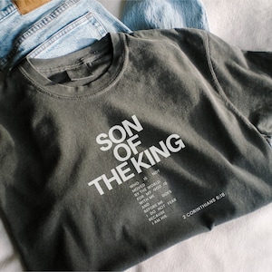 May include: A charcoal gray crew neck t-shirt featuring white text that reads SON OF THE KING, followed by a verse that says WHO IS NOT MOVED BY THE WORLD, FOR MY GOD IS WITH ME AND BEFORE ME. I DO NOT FEAR BECAUSE I AM HIS. 2 CORINTHIANS 6:18.