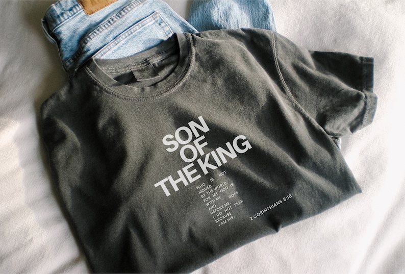 May include: A charcoal gray crew neck t-shirt featuring white text that reads SON OF THE KING, followed by a verse that says WHO IS NOT MOVED BY THE WORLD, FOR MY GOD IS WITH ME AND BEFORE ME. I DO NOT FEAR BECAUSE I AM HIS. 2 CORINTHIANS 6:18.