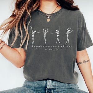 May include: A gray t-shirt with a graphic of five skeletons dancing. The text "dry bones come alive" and "Ezekiel 37:5" is printed below the skeletons.
