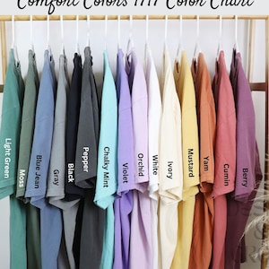 K&ouml;nnte beinhalten: Ein Gestell mit 12 T-Shirts in verschiedenen Farben, darunter Hellgr&uuml;n, Moos, Jeansblau, Grau, Schwarz, Pfeffer, Kreideminze, Violett, Orchidee, Wei&szlig;, Elfenbein, Senf, S&uuml;&szlig;kartoffel, Kreuzk&uuml;mmel und Beere.