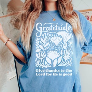 May include: A blue short-sleeve t-shirt featuring a white graphic print. The design includes the word Gratitude at the top, a central illustration of various stylized flowers and leaves, and the text Give thanks to the Lord for He is good.