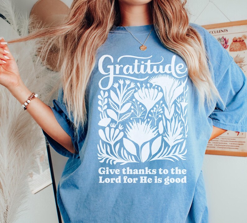 May include: A blue short-sleeve t-shirt featuring a white graphic print. The design includes the word Gratitude at the top, a central illustration of various stylized flowers and leaves, and the text Give thanks to the Lord for He is good.