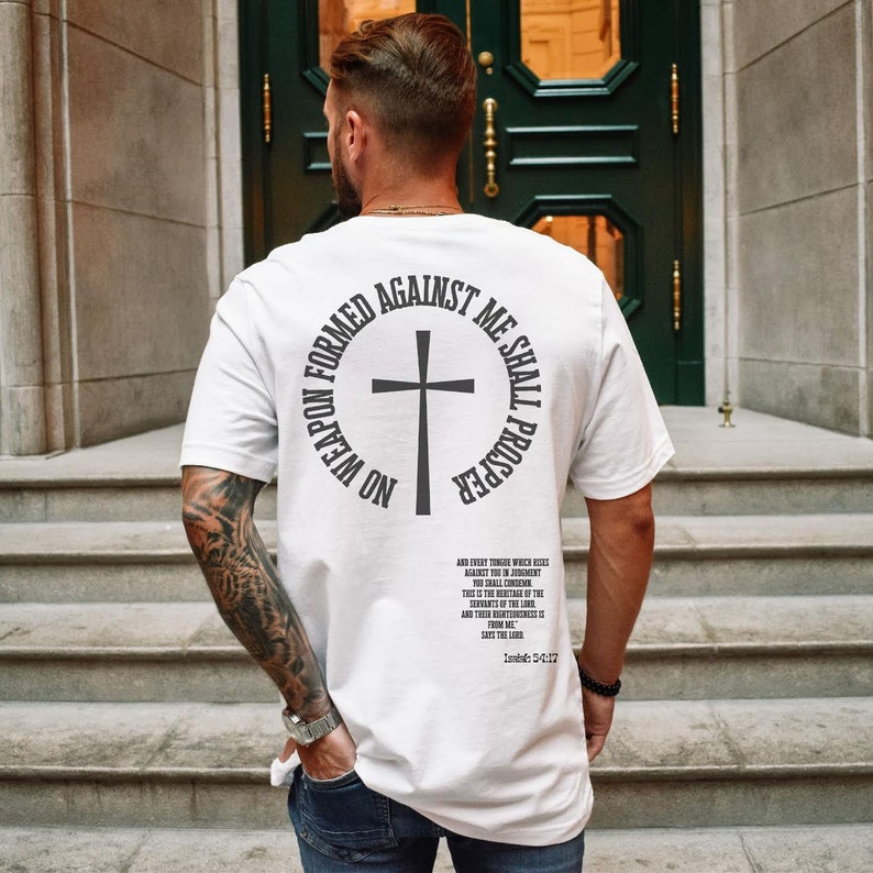Pode incluir: Camiseta branca com um gr&aacute;fico preto nas costas. O gr&aacute;fico &eacute; um c&iacute;rculo com uma cruz no centro e o texto "No weapon formed against me shall prosper". O texto est&aacute; em negrito, sem serifa. H&aacute; tamb&eacute;m um texto menor abaixo do gr&aacute;fico, que &eacute; um vers&iacute;culo b&iacute;blico.