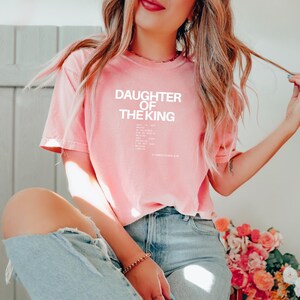 May include: A pink t-shirt with the text "Daughter of the King" and a Bible verse printed on the front.
