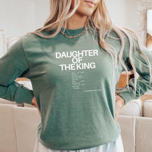 May include: A green long-sleeved sweatshirt with the text "Daughter of the King" in white. The text continues with a quote from 2 Corinthians 6:18, "Who is not moved by the world, for the world is not worthy of my God. He is with me, and He is before me. He does not flap because I fear Him. I am His."