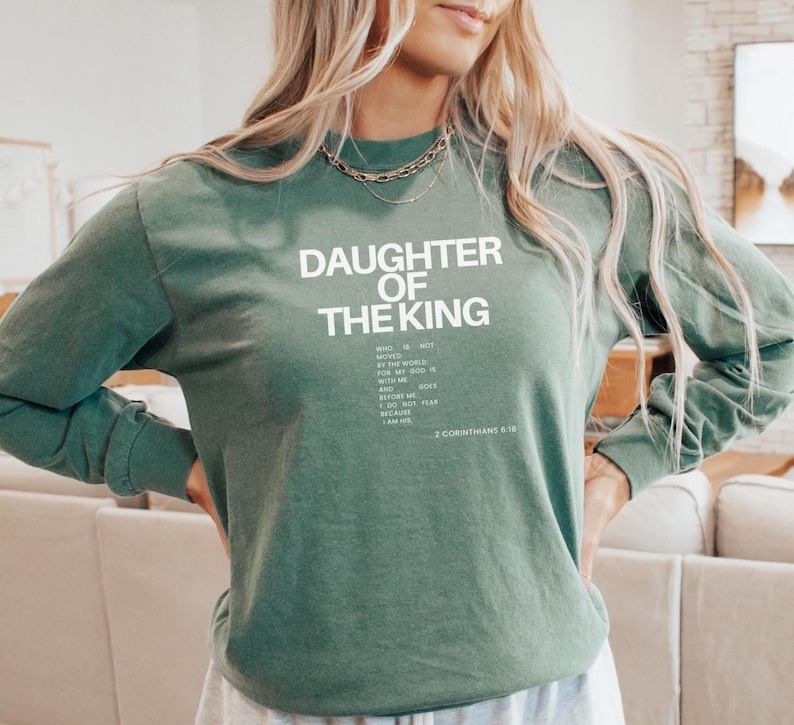 May include: A green long-sleeved sweatshirt with the text "Daughter of the King" in white. The text continues with a quote from 2 Corinthians 6:18, "Who is not moved by the world, for the world is not worthy of my God. He is with me, and He is before me. He does not flap because I fear Him. I am His."