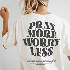 Puede incluir: Camiseta blanca con texto negro que dice "Reza m&aacute;s, preoc&uacute;pate menos" y una cita de Filipenses 4:6-7.