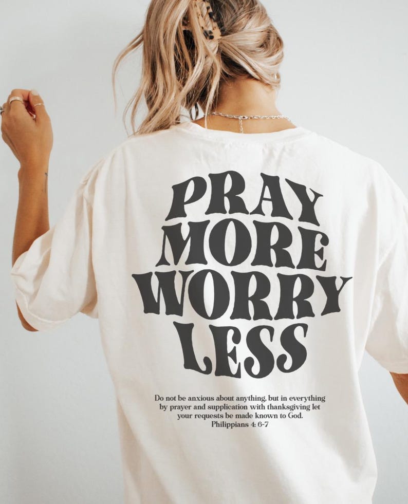 Puede incluir: Camiseta blanca con texto negro que dice "Reza m&aacute;s, preoc&uacute;pate menos" y una cita de Filipenses 4:6-7.
