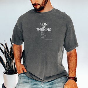 May include: A charcoal gray short-sleeve t-shirt featuring the white text SON OF THE KING followed by smaller text and the citation 2 CORINTHIANS 5:20. The shirt has a crew neckline and a relaxed fit with a slightly faded, washed-out fabric appearance.