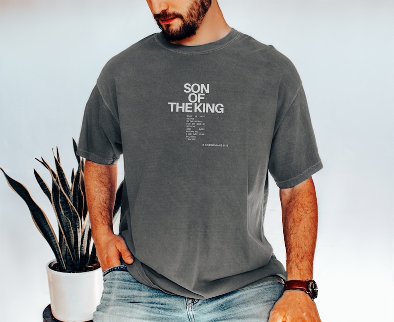May include: A charcoal gray short-sleeve t-shirt featuring the white text SON OF THE KING followed by smaller text and the citation 2 CORINTHIANS 5:20. The shirt has a crew neckline and a relaxed fit with a slightly faded, washed-out fabric appearance.