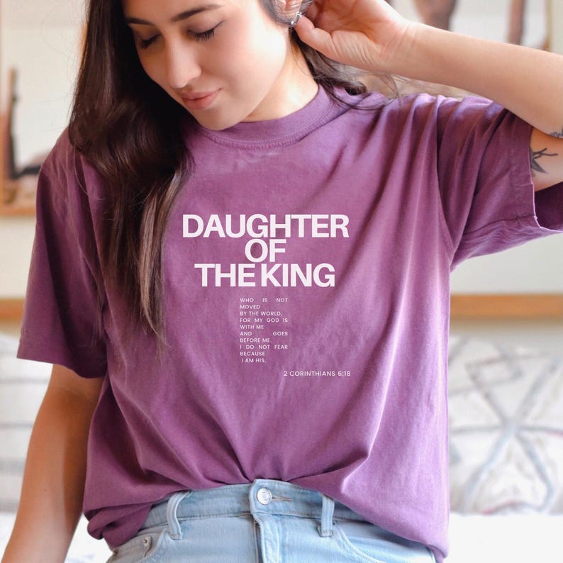 May include: Purple t-shirt with the text "Daughter of the King" and a Bible verse from 2 Corinthians 6:18. The verse reads: "Who is not moved by the world, for my God is with me and before me goes. I do not fear because I am His."