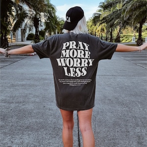 Puede incluir: Una camiseta negra con texto blanco que dice "Reza m&aacute;s, preoc&uacute;pate menos". El texto est&aacute; en un estilo de fuente retro. Debajo del texto hay una cita de Filipenses 4:6-7.